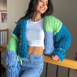 May include: A chunky knit cardigan in shades of blue and green. The oversized sweater features large, visible stitches and a cropped design. The sleeves are a mix of light blue, teal, and lime green. The cardigan is worn with a white top and blue jeans.