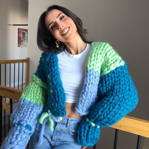 May include: A chunky knit cardigan in shades of teal, light blue, and green. The oversized sweater features a tie closure and is made of thick, soft yarn. The sleeves are a prominent feature, adding to the cozy, handmade aesthetic.