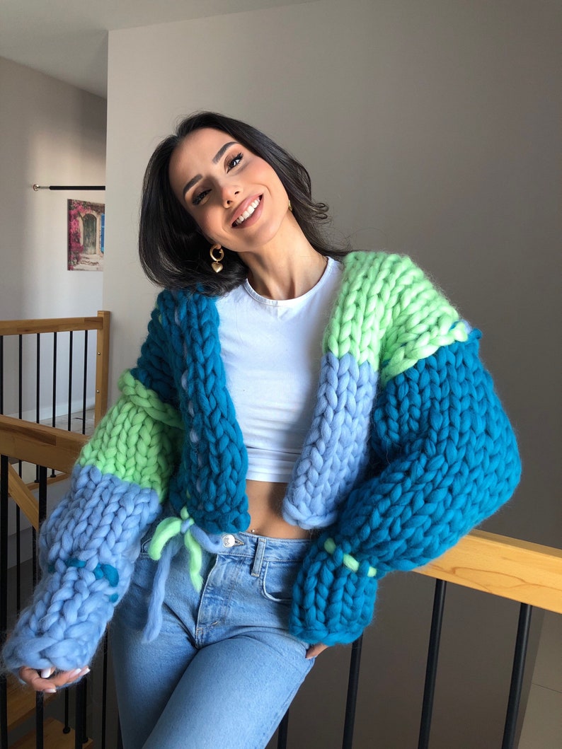May include: A chunky knit cardigan in shades of teal, light blue, and green. The oversized sweater features a tie closure and is made of thick, soft yarn. The sleeves are a prominent feature, adding to the cozy, handmade aesthetic.