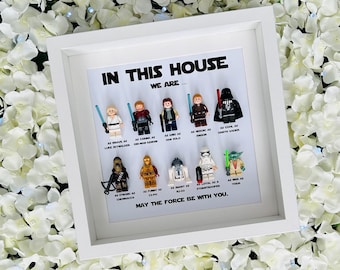 May include: A white square shadow box frame containing ten Star Wars character minifigures arranged in two rows. The text reads: IN THIS HOUSE WE ARE... followed by character names and traits, ending with MAY THE FORCE BE WITH YOU. The background is white.