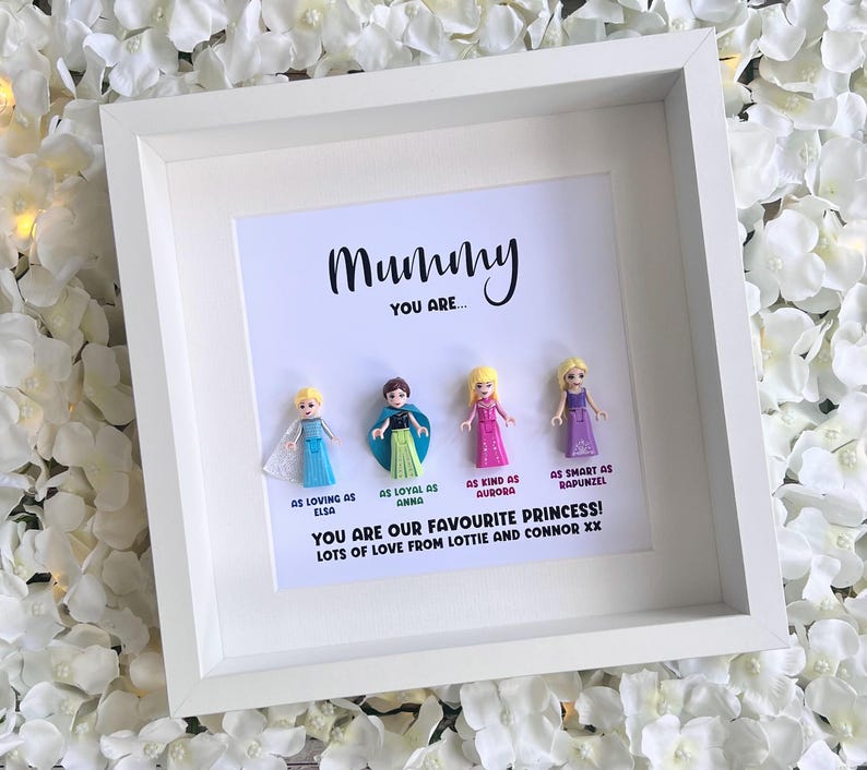 May include: A white frame with a white mat and a white background. The frame contains a message that reads "Mummy You Are..." and four small plastic figurines of Disney princesses. The text below the figurines reads "As loving as Elsa, As loyal as Anna, As kind as Aurora, As smart as Rapunzel. You are our favourite princess! Lots of love from Lottie and Connor xx."