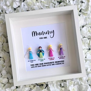 May include: A white picture frame with a white background and a black inscription that reads "Mummy You Are..."  The frame contains five small plastic figurines of Disney princesses.  The inscription below the figurines reads "As loving as Elsa, As loyal as Anna, As kind as Aurora, As smart as Rapunzel. You are our favourite princess! Lots of love from Lottie and Connor xx"