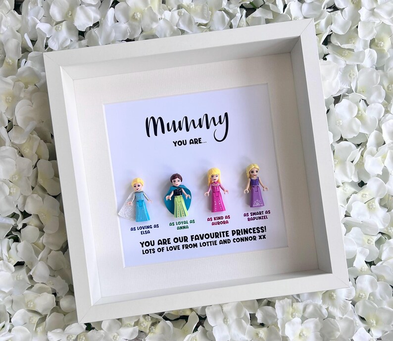 May include: A white picture frame with a white background and a black inscription that reads "Mummy You Are..."  The frame contains five small plastic figurines of Disney princesses.  The inscription below the figurines reads "As loving as Elsa, As loyal as Anna, As kind as Aurora, As smart as Rapunzel. You are our favourite princess! Lots of love from Lottie and Connor xx"