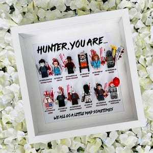 May include: A white shadow box frame containing twelve horror-themed minifigures arranged in two rows. The text reads HUNTER, YOU ARE... and WE ALL GO A LITTLE MAD SOMETIMES, with individual character names and traits printed below each figure.