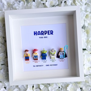 Op de afbeelding: Een wit frame met een witte passe-partout en een witte achtergrond. De achtergrond heeft de tekst "HARPER YOU ARE..." en toont vijf speelgoedfiguren uit de Toy Story-franchise. De figuren zijn Woody, Jessie, Alien, Buzz Lightyear en Bo Peep. De tekst onder de figuren luidt "AS LOYAL AS WOODY, AS FRIENDLY AS JESSIE, AS COOL AS ALIEN, AS BRAVE AS BUZZ, AS CHARMING AS BO PEEP. TO INFINITY... AND BEYOND!"