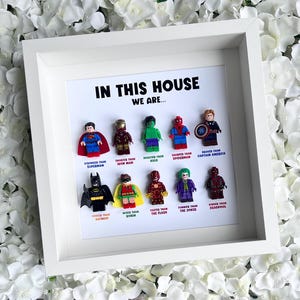 May include: A white framed print featuring the text "IN THIS HOUSE WE ARE..." and Lego figures of superheroes. The characters include Superman, Iron Man, Hulk, Spider-Man, Captain America, Batman, Robin, The Flash, The Joker, and Deadpool.