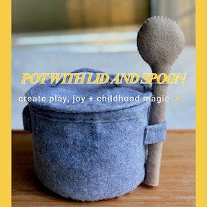 Puede incluir: Una olla de fieltro con tapa y cuchara. La olla es de color azul grisáceo apagado, con una tapa a juego y dos asas pequeñas. La cuchara es de color beige claro y está sujeta a la olla con un lazo de tela. El texto "POT WITH LID AND SPOON" se muestra en amarillo.
