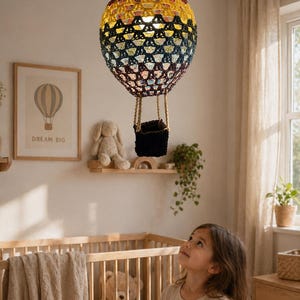 May include: A colorful, crocheted hot air balloon lamp hangs from the ceiling. The lamp features a basket and is suspended above a crib. The balloon is a mix of colors, including yellow, blue, and burgundy. A child looks up at the lamp.