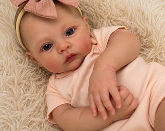 May include: A realistic baby doll with blue eyes and fair skin, wearing a light peach-coloured short-sleeved onesie. The doll features a textured dusty rose fabric bow headband positioned on its head, with a soft, lifelike facial expression.