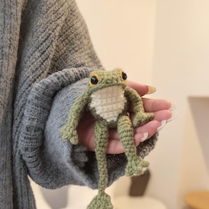 Puede incluir: Una muñeca de rana de ganchillo hecha a mano en tonos verdes y crema. La rana tiene ojos negros grandes, una barriga blanca y pies detallados. La muñeca se sostiene en una mano, mostrando el intrincado trabajo de ganchillo.