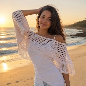 Peut inclure: Un haut en crochet blanc avec des franges. Le haut a un design épaules nues et un tissage lâche et ouvert. Le modèle porte le haut sur une plage au coucher du soleil. Le haut fait partie d'une collection de vêtements de plage.