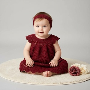Peut inclure: Un bébé porte une robe et un bandeau en crochet bordeaux, assis sur un tapis crème. La robe présente un ourlet festonné et des découpes florales. Des roses artificielles bordeaux et crème sont placées à côté du bébé.