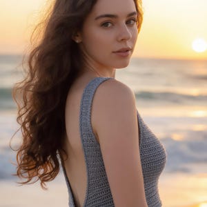 Puede incluir: Un top de ganchillo gris con espalda baja y detalle de lazo. El top es sin mangas y parece hecho a mano. El fondo muestra una escena de playa con una puesta de sol.