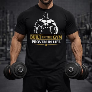 以下が含まれることがあります： 白い筋肉質な人物のグラフィックと「BUILT IN THE GYM PROVEN IN LIFE」のテキストが入った黒いTシャツ。ジムで、その人は2つの黒いダンベルを持っています。