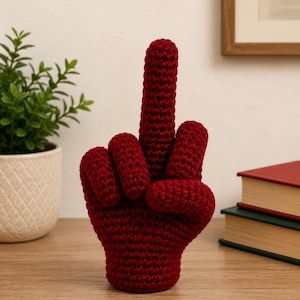 Puede incluir: Una escultura de mano tejida a ganchillo en color rojo oscuro que muestra un gesto de dedo medio. El objeto está elaborado con hilo con un patrón de puntos texturizado visible, creando una forma firme y vertical que se sostiene sola.