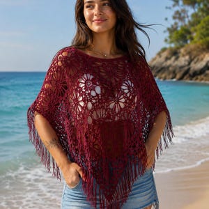 Peut inclure: Un poncho en crochet bordeaux avec un motif floral et des franges. Le haut en maille ajourée est porté sur un haut blanc et un short en jean. Le cadre de la plage présente une eau bleue et un ciel clair.