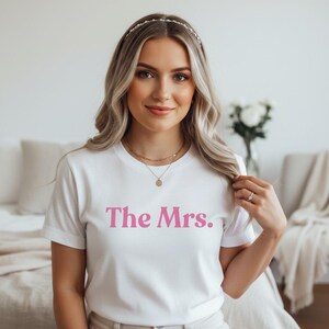 Peut inclure: T-shirt blanc avec l'inscription "The Mrs." en rose. Le t-shirt est de style col rond et semble être fait d'une matière douce. Le texte est centré sur le devant du t-shirt.