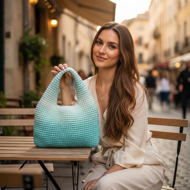 May include: A crocheted tote bag with a gradient of light blue to teal. The bag has a rounded shape and a wide handle. The bag is sitting on a wooden table.