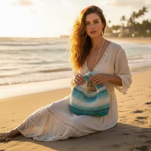 May include: A crocheted handbag with a round handle, featuring horizontal stripes of white, light blue, and teal. The bag has a tassel and is held against a beach backdrop. The overall aesthetic is casual and beachy.