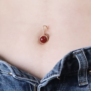 Op de afbeelding: Gouden navelring met een rode edelsteen. De ring is haakvormig.