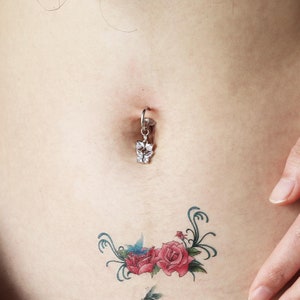 Puede incluir: Primer plano del abdomen de una persona con un piercing en el ombligo con un dije de mariposa plateado. Un pequeño tatuaje floral es visible debajo del piercing.