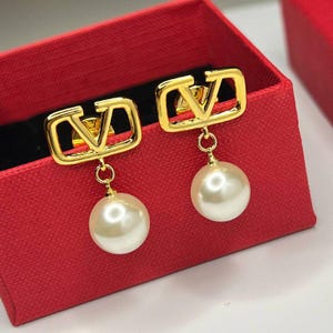 May include: Gold-tone earrings featuring a square frame with a 'V' design, suspending a large, round, off-white pearl. The earrings are displayed inside a red box, showcasing the jewelry's elegant design.