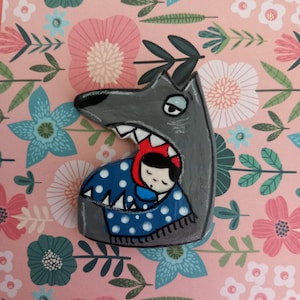 Peut inclure: Une broche en bois peinte à la main représentant un loup gris à la gueule ouverte, révélant un petit personnage portant une capuche rouge et enveloppé dans une couverture bleue à pois blancs. Le loup a des dents blanches acérées.