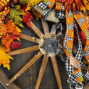May include: A rustic wooden wagon wheel decorated with fall foliage, sunflowers, and pumpkin-themed ribbons. The arrangement includes orange, yellow, and red leaves, berries, and decorative gourds, creating a festive autumn theme.