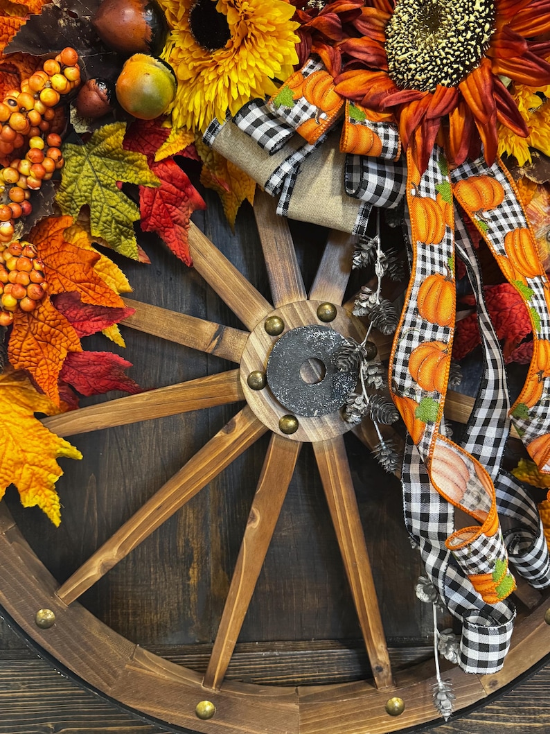 May include: A rustic wooden wagon wheel decorated with fall foliage, sunflowers, and pumpkin-themed ribbons. The arrangement includes orange, yellow, and red leaves, berries, and decorative gourds, creating a festive autumn theme.