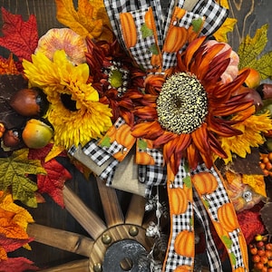 May include: A fall-themed wreath featuring sunflowers, pumpkins, and autumn leaves. The wreath includes a wooden wagon wheel and is decorated with a black and white checkered ribbon with orange pumpkin accents. The color palette includes shades of orange, yellow, and red.