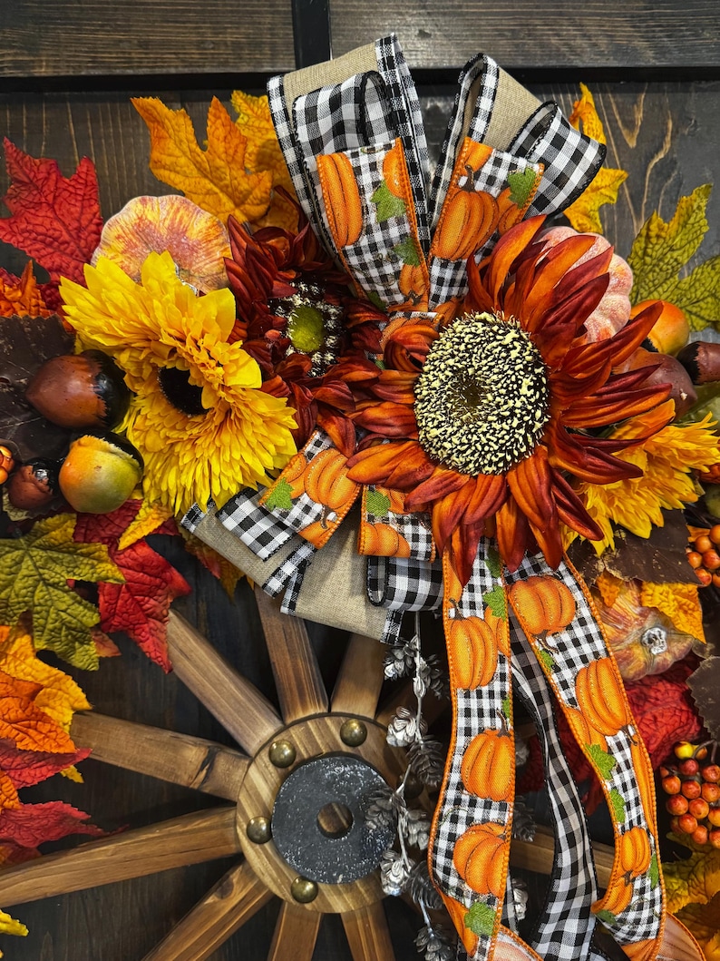 May include: A fall-themed wreath featuring sunflowers, pumpkins, and autumn leaves. The wreath includes a wooden wagon wheel and is decorated with a black and white checkered ribbon with orange pumpkin accents. The color palette includes shades of orange, yellow, and red.