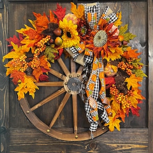 May include: A fall-themed wreath featuring a wooden wagon wheel base. The wreath is adorned with vibrant orange, yellow, and red leaves, sunflowers, berries, and pumpkins. A black and white checkered ribbon with pumpkin accents adds a decorative touch.