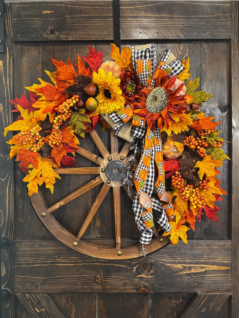 May include: A fall-themed wreath featuring a wooden wagon wheel base. The wreath is adorned with vibrant orange, yellow, and red leaves, sunflowers, berries, and pumpkins. A black and white checkered ribbon with pumpkin accents adds a decorative touch.