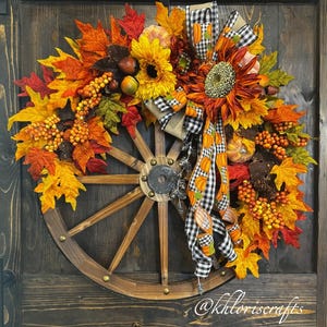 May include: A rustic fall wreath featuring a wooden wagon wheel base. The wreath is adorned with vibrant autumn leaves in shades of red, orange, and yellow, sunflowers, berries, and a decorative bow with pumpkin accents.