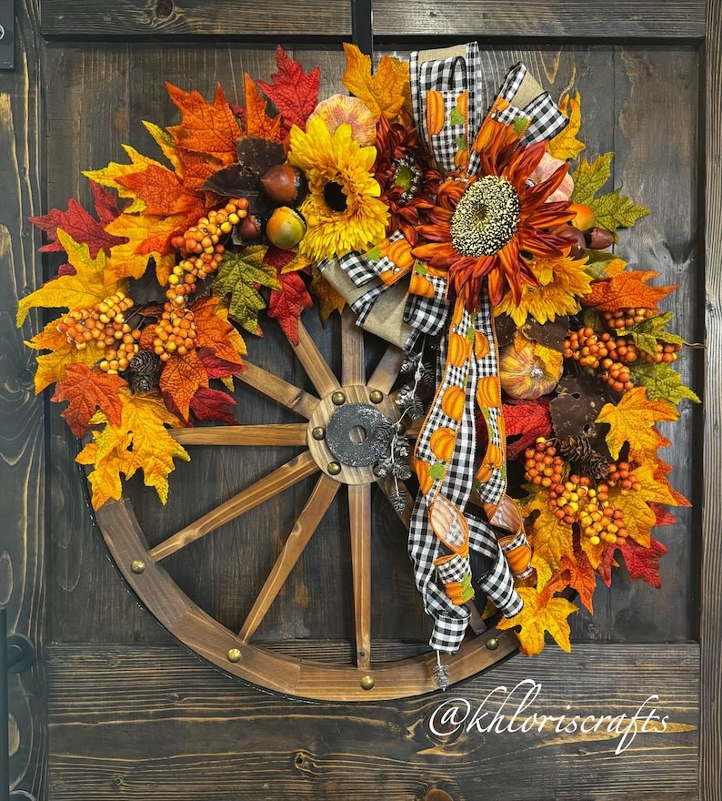 May include: A rustic fall wreath featuring a wooden wagon wheel base. The wreath is adorned with vibrant autumn leaves in shades of red, orange, and yellow, sunflowers, berries, and a decorative bow with pumpkin accents.