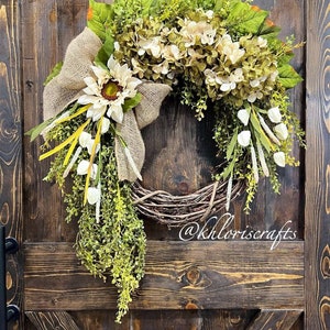 May include: A wreath made of grapevine with a burlap bow and white hydrangeas. The wreath is decorated with green leaves and white flowers. The wreath is hanging on a wooden door with the text "@khloriscrafts" written on the door.