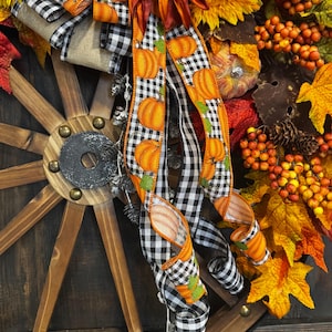 May include: A fall-themed wreath featuring a wooden wagon wheel base. The wreath is adorned with sunflowers, orange berries, and autumn leaves. A black and white checkered ribbon with orange pumpkin accents adds a festive touch.