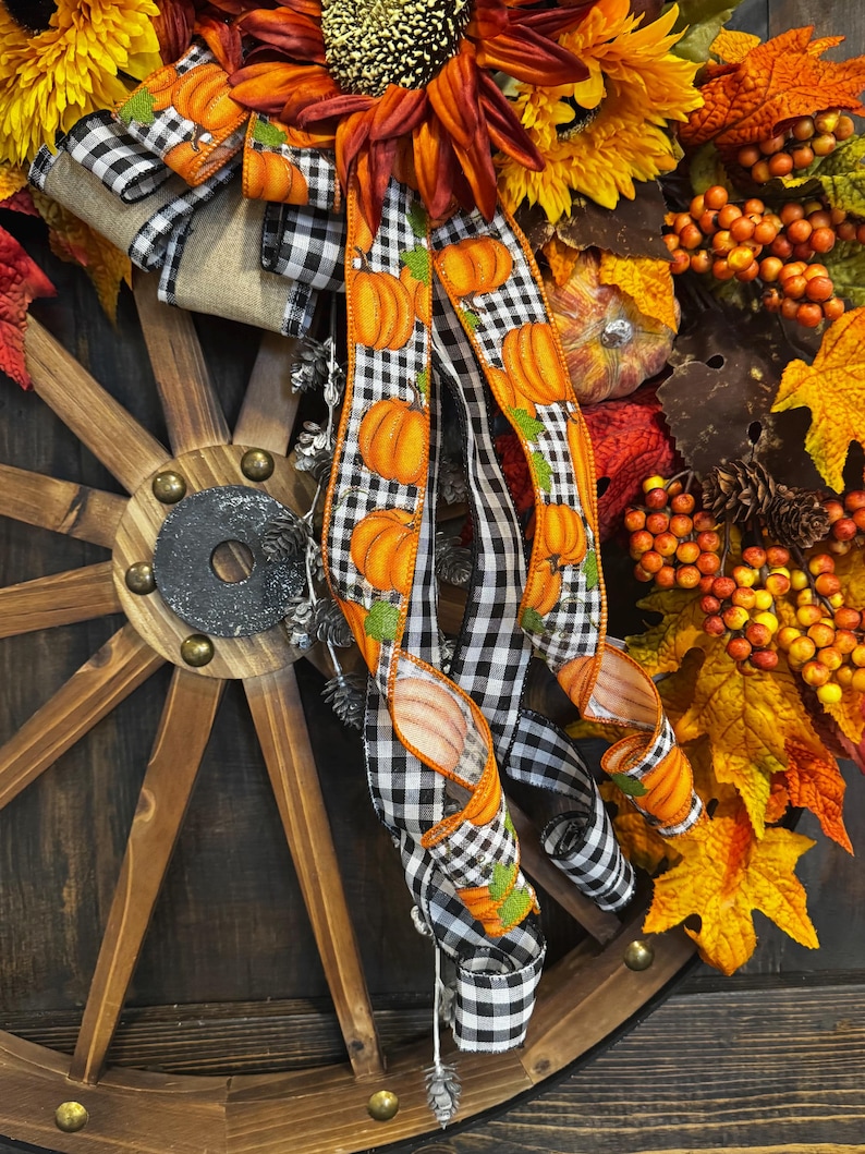 May include: A fall-themed wreath featuring a wooden wagon wheel base. The wreath is adorned with sunflowers, orange berries, and autumn leaves. A black and white checkered ribbon with orange pumpkin accents adds a festive touch.