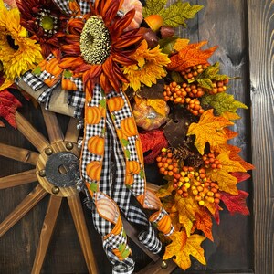 May include: A fall-themed wreath featuring a wooden wagon wheel base. The wreath is adorned with sunflowers, pumpkins, fall leaves, and berries. A black and white checkered ribbon with pumpkin accents adds a decorative touch. The color palette includes orange, yellow, and red.