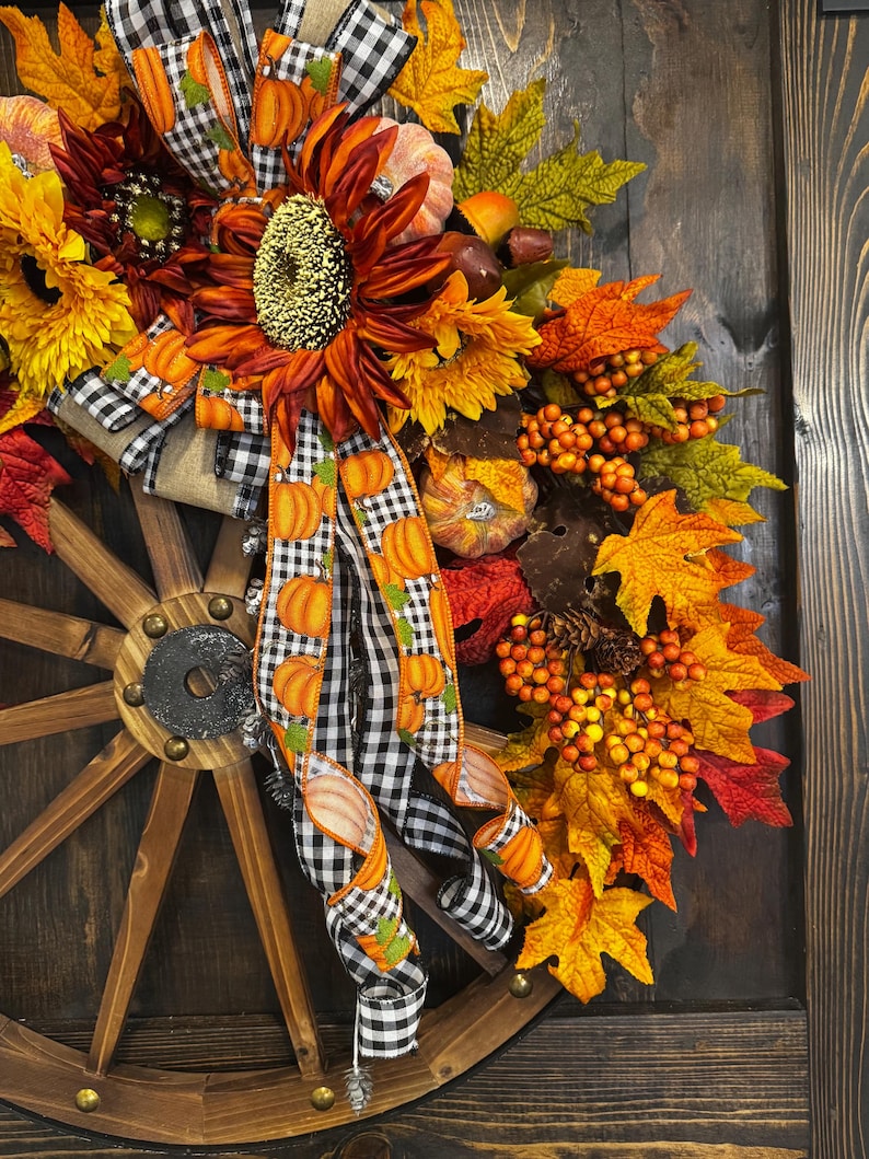 May include: A fall-themed wreath featuring a wooden wagon wheel base. The wreath is adorned with sunflowers, pumpkins, fall leaves, and berries. A black and white checkered ribbon with pumpkin accents adds a decorative touch. The color palette includes orange, yellow, and red.
