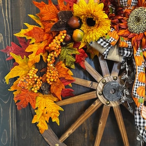 May include: A rustic fall-themed wagon wheel wreath. It features vibrant sunflowers, colorful autumn leaves in shades of red, orange, and yellow, and decorative berries. The wreath is adorned with a black and white checkered ribbon and pumpkin accents.