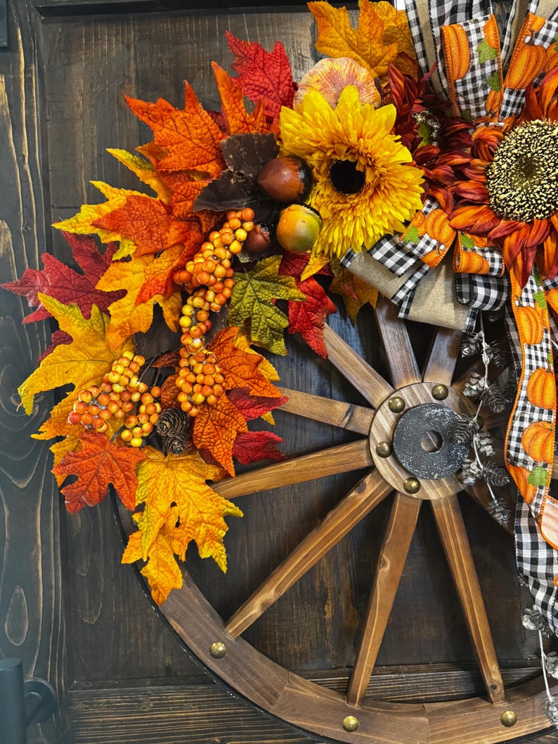 May include: A rustic fall-themed wagon wheel wreath. It features vibrant sunflowers, colorful autumn leaves in shades of red, orange, and yellow, and decorative berries. The wreath is adorned with a black and white checkered ribbon and pumpkin accents.