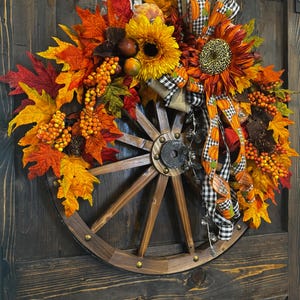 May include: A rustic fall wreath featuring a wooden wagon wheel base adorned with vibrant autumn leaves in shades of red, orange, and yellow. Sunflowers, berries, and decorative ribbons add to the seasonal charm. The wreath is mounted on a dark wooden door.