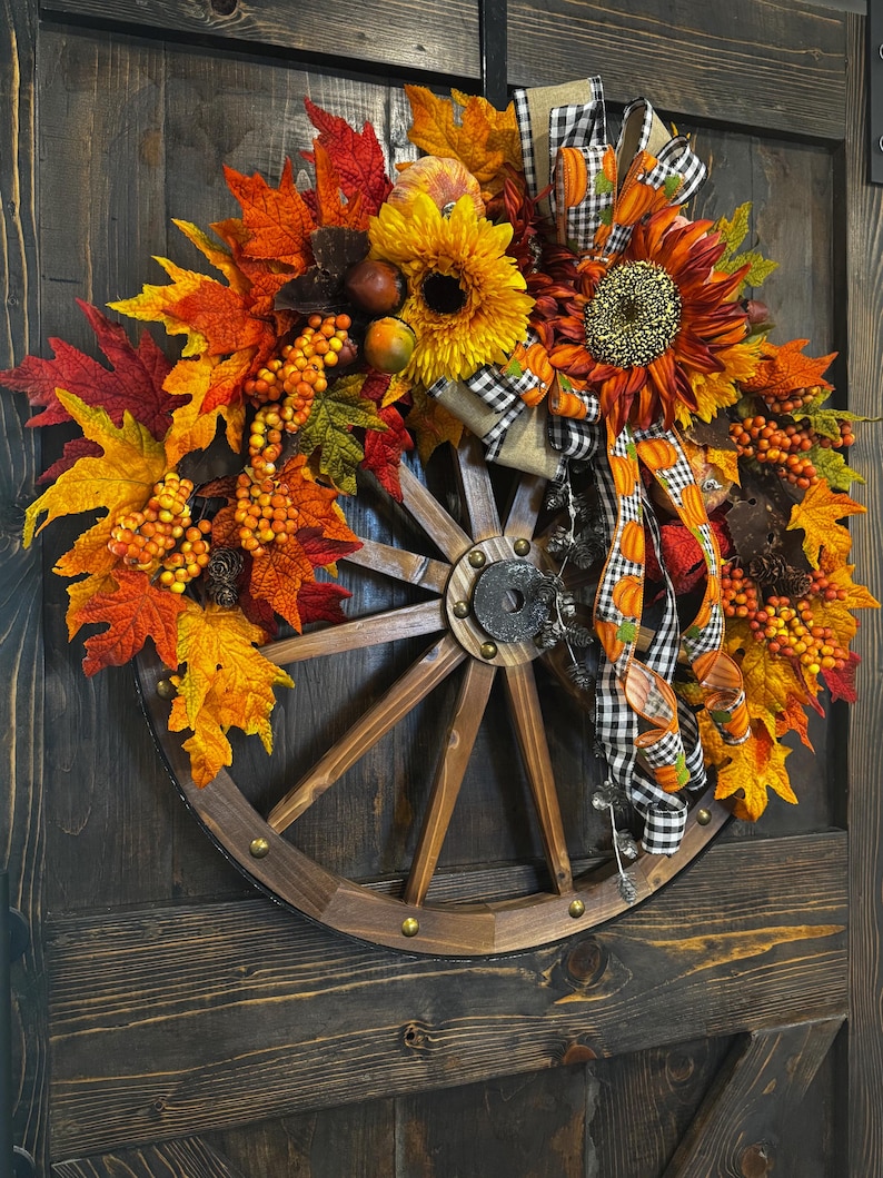 May include: A rustic fall wreath featuring a wooden wagon wheel base adorned with vibrant autumn leaves in shades of red, orange, and yellow. Sunflowers, berries, and decorative ribbons add to the seasonal charm. The wreath is mounted on a dark wooden door.