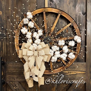 May include: A rustic wreath made with a wooden wagon wheel, white cotton bolls, and a beige and white polka dot bow. The wreath is hanging on a dark brown wooden door with the text "@khloriscrafts" at the bottom.