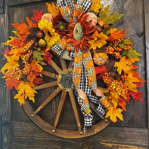 May include: A fall-themed wreath featuring a wooden wagon wheel base. The wreath is adorned with sunflowers, pumpkins, and colorful autumn leaves in shades of orange, yellow, and red. A black and white checkered ribbon with pumpkin accents adds a decorative touch.