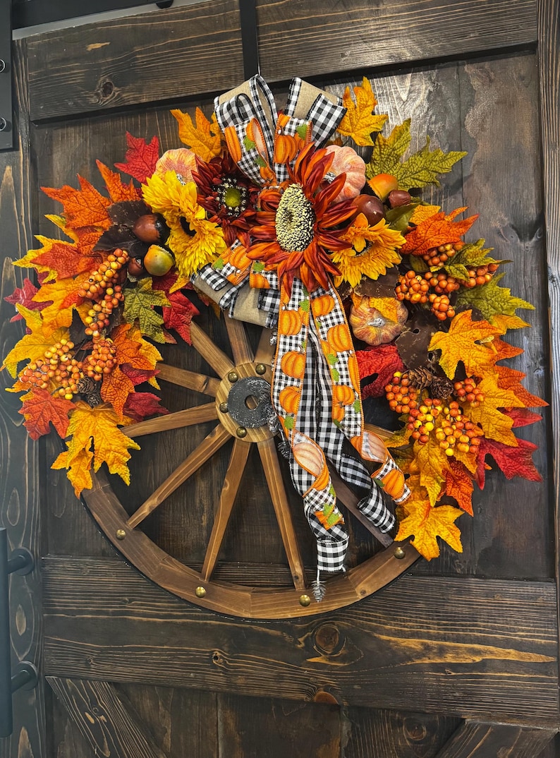 May include: A fall-themed wreath featuring a wooden wagon wheel base. The wreath is adorned with sunflowers, pumpkins, and colorful autumn leaves in shades of orange, yellow, and red. A black and white checkered ribbon with pumpkin accents adds a decorative touch.