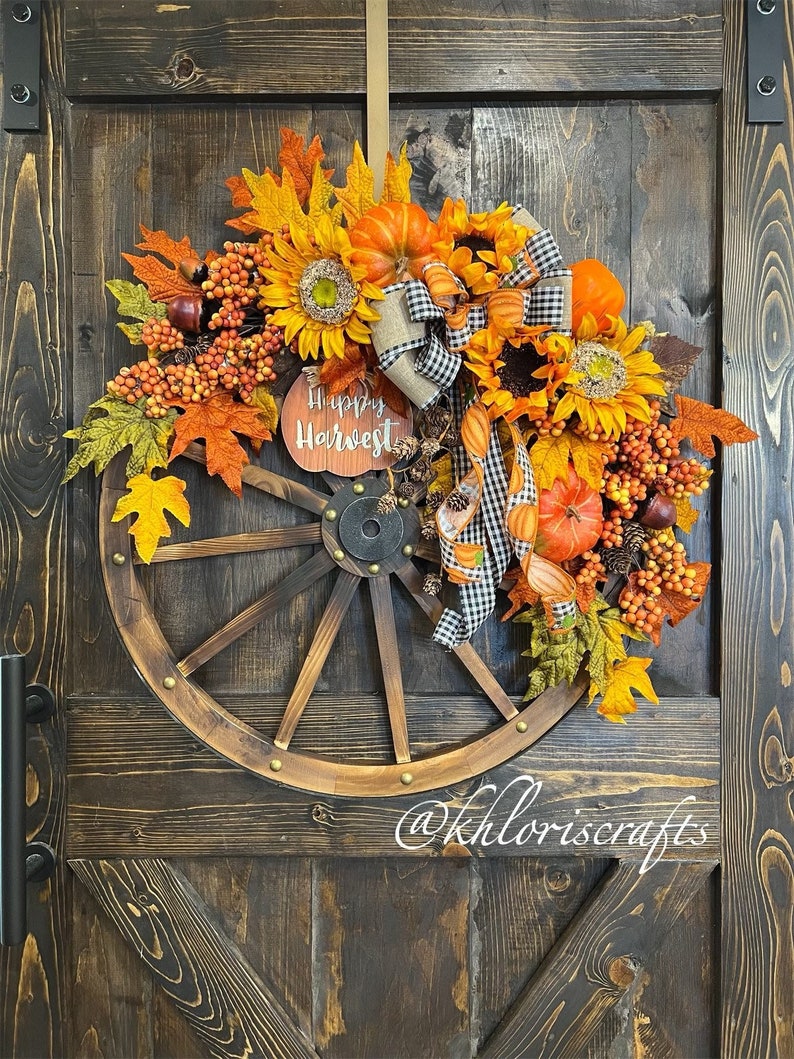 May include: A fall-themed wreath with a wooden wagon wheel base. The wreath is decorated with sunflowers, fall leaves, pumpkins, berries, and a sign that reads 'Happy Harvest'. The wreath is hanging on a wooden door.