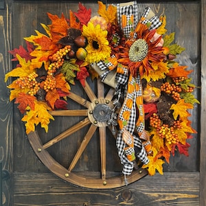 May include: A rustic fall wreath featuring a wooden wagon wheel base. The wreath is adorned with vibrant autumn leaves in shades of red, orange, and yellow, sunflowers, berries, and a black and white checkered bow.