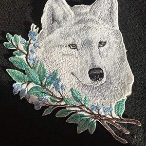 Puede incluir: Parche bordado para planchar con un lobo blanco con bayas azules y hojas verdes.
